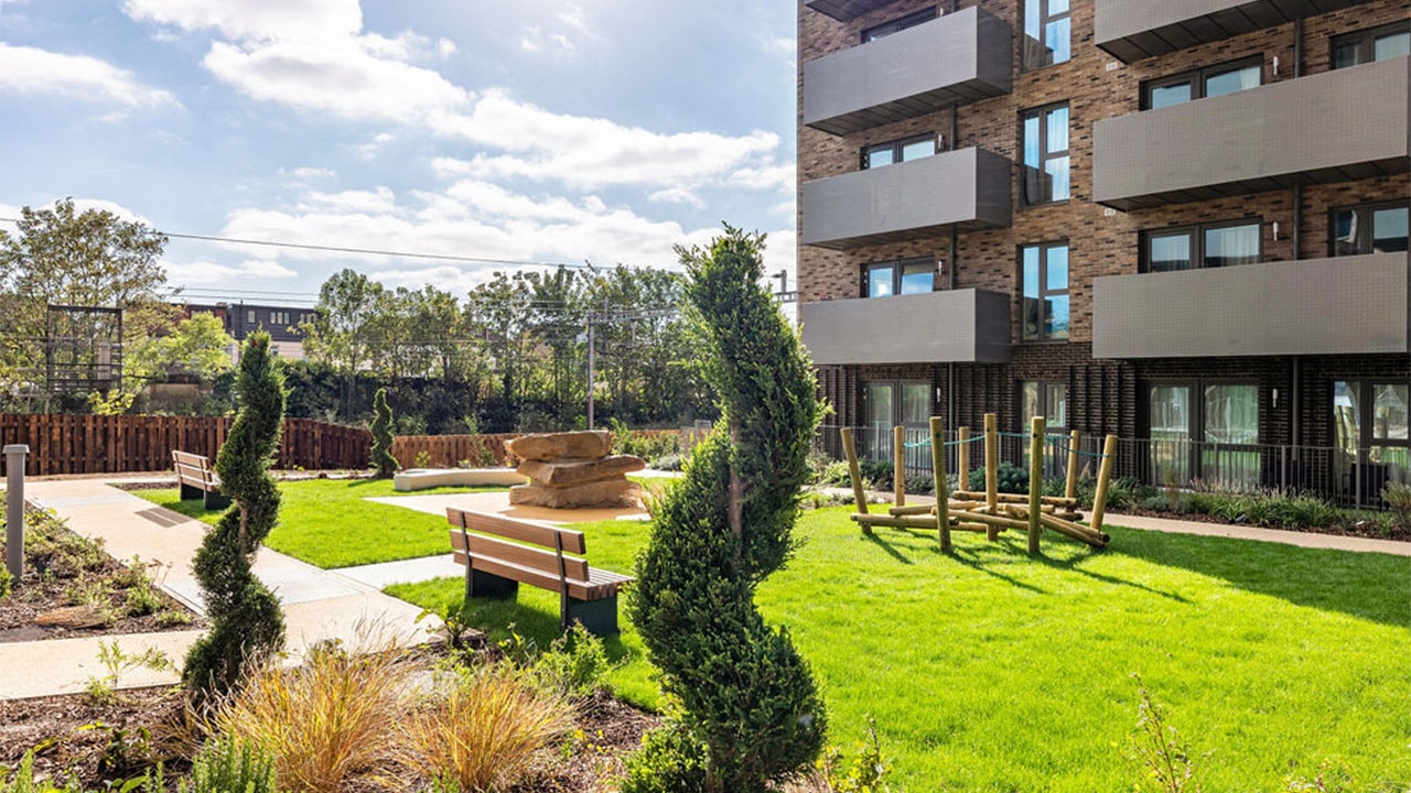 Landscaped communal garden at the One Goodmayes development, featuring lawns, planting beds, seating areas, pathways and outdoor play equipment.
